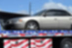 A silver car on a patriotic-themed tow truck with "Lockout," "Jumpstart," "24 Hour" text. Clear skies and building in the background.