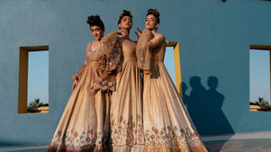 Three women in ornate gold gowns pose against a blue wall with window cutouts, casting long shadows in warm sunlight.
