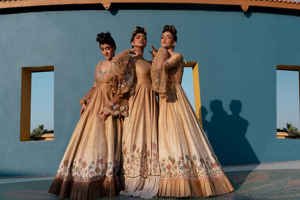 Three women in ornate gold gowns pose against a blue wall with window cutouts, casting long shadows in warm sunlight.