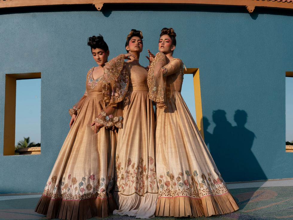 Three women in ornate gold gowns pose against a blue wall with window cutouts, casting long shadows in warm sunlight.