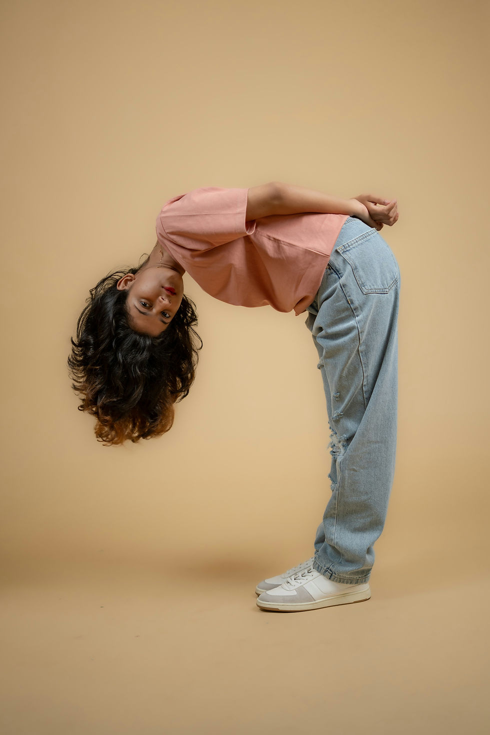 Person bends forward, looking at the camera, in pink top and jeans. Beige backdrop sets a curious, playful mood.