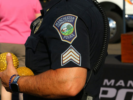 Police officer in a dark uniform with "Manchester Connecticut Police" patch holds two yellow balls. Background with people and vehicle.
