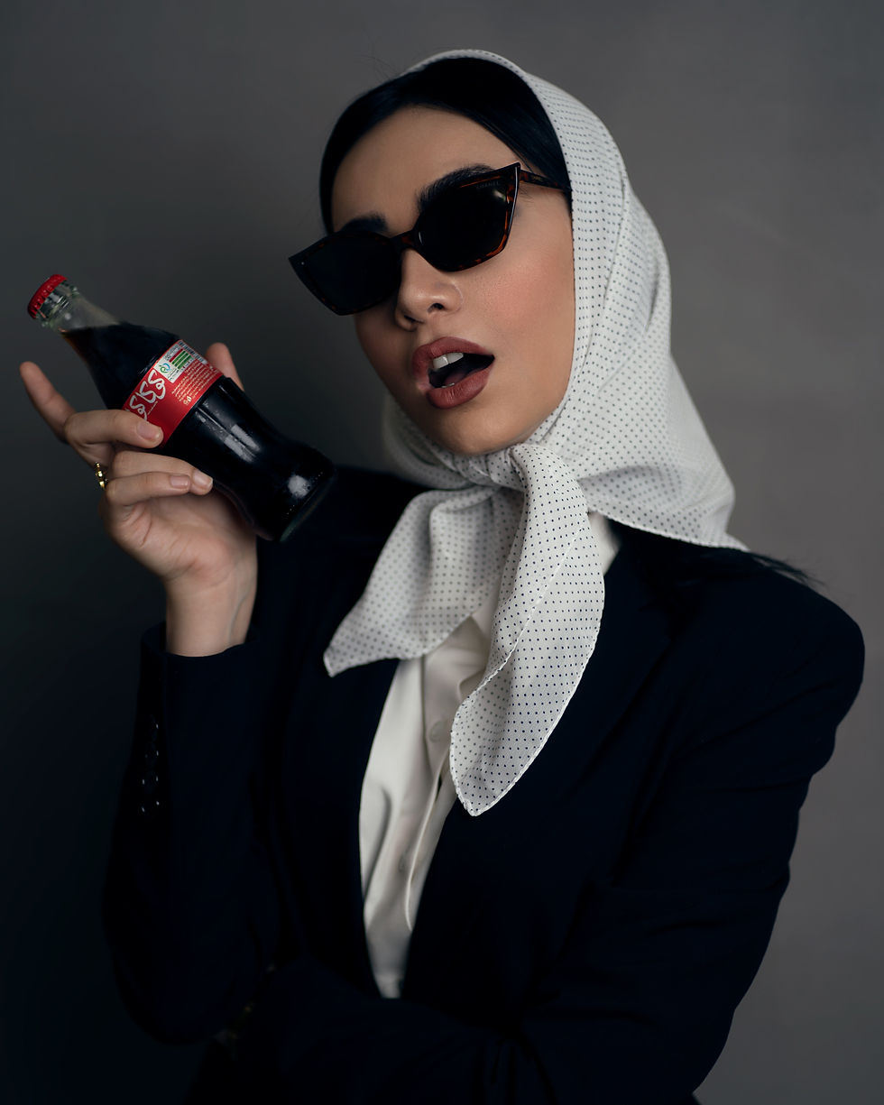 Woman in sunglasses and polka-dot headscarf holding a soda bottle labeled "555," against a gray background, looking surprised.