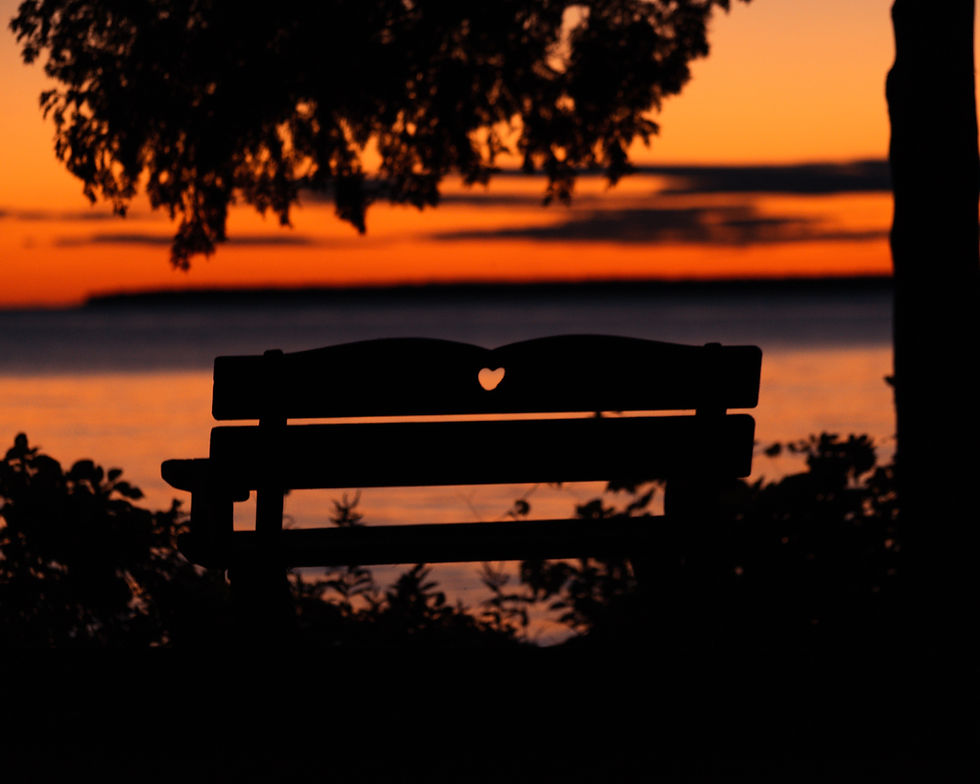 Silhouette of a bench with heart cutout at sunset, trees framing the glowing orange sky and calm lake in the background, peaceful mood.