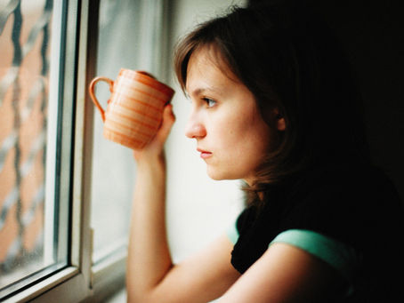 Woman with brown hair holding an orange striped mug, gazes pensively out a window. The mood is contemplative.
