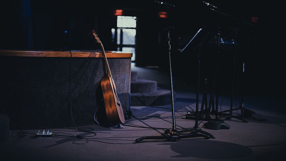 Acoustic guitar leans against a stage podium with microphones and cables in a dimly lit room. Bright exit signs are visible in the background.