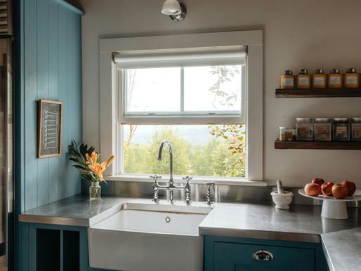 Bright kitchen sink under window, teal cabinets, shelf of labeled jars, apples on a pedestal, and orange lilies by a chalkboard.
