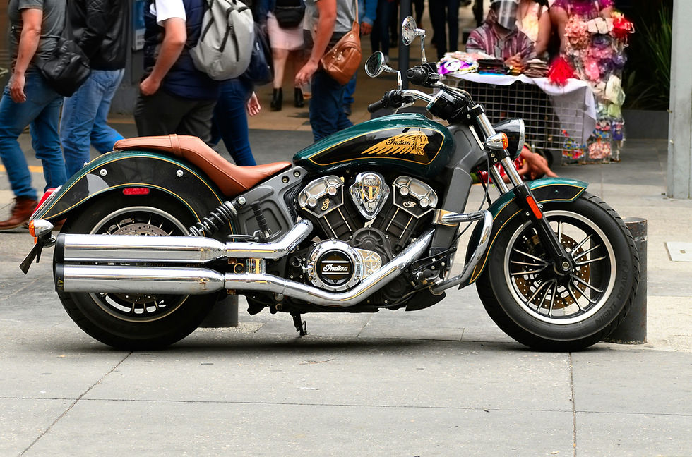 A parked Indian motorcycle with chrome pipes and brown seat stands on a busy street. Pedestrians walk by, and a nearby colorful market stall adds vibrancy.