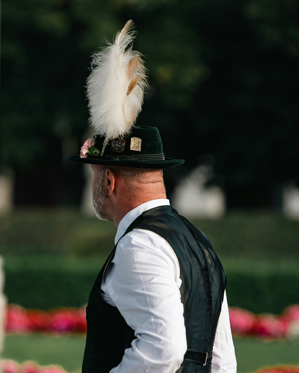 Man in traditional black hat with tall white feather, seen from behind at an outdoor event with blurred flowers and greenery.