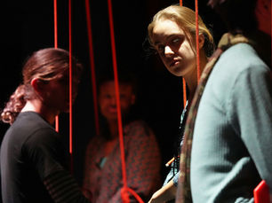 Two people framed by red cords resembling puppet strings, creating an unsettling sense of control, conformity, and performance in the workplace.