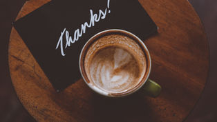 Latte with latte art in green mug on wooden table, beside a black card with "Thanks!" written in white. Cozy and appreciative mood.