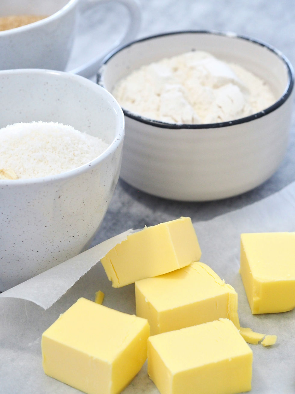 Bowls of flour and sugar next to cubes of yellow butter on parchment paper, set on a gray counter. Bright and inviting baking scene.