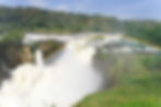 A breathtaking view of Murchison Falls, where the Nile River thunders through a narrow gorge, accompanied by a rainbow and lush greenery.