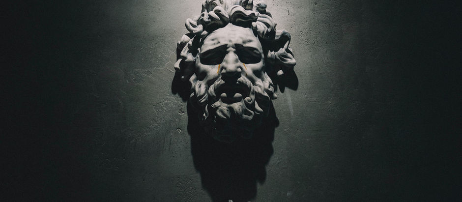 Three stone heads illuminated on a dark wall. The top face is serene, middle is expressive, and bottom is obscured in shadows. Dramatic lighting.