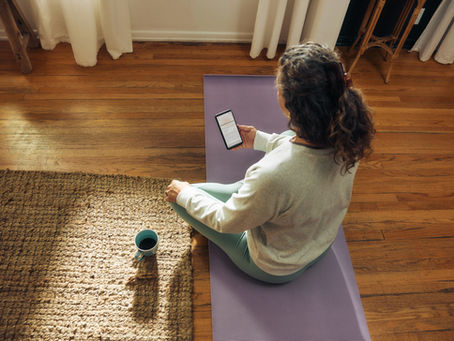 Woman with wellness routine sitting on a purple yoga mat, holding a phone, in a sunlit room with wooden floors. A cup of coffee is on a woven rug nearby.