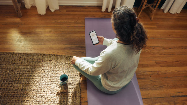 Person with long, curly brown hair sitting on a purple yoga mat with a smartphone in one hand and a mug next to them, representing an always-on world and setting boundaries around technology