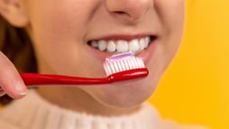 A person smiling with a red toothbrush and pink toothpaste near their teeth, set against a bright yellow background.