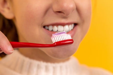 A person smiling with a red toothbrush and pink toothpaste near their teeth, set against a bright yellow background.