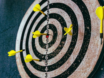 Yellow darts on a circular dartboard with numbers, three near the bullseye. Darker background adds contrast, emphasizing focus and precision.
