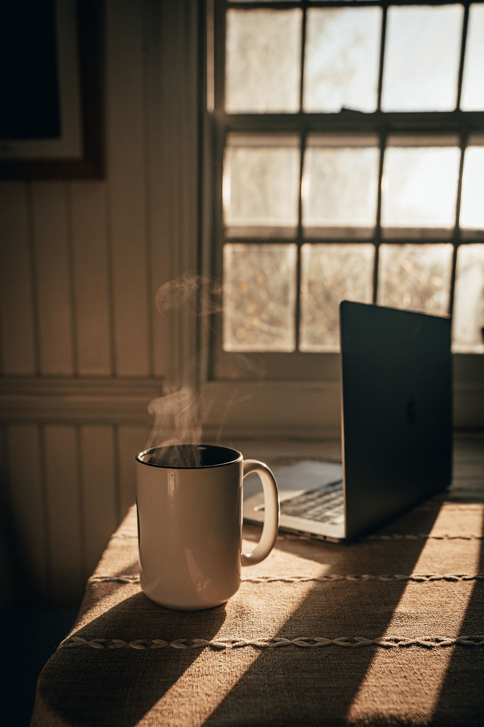 A steaming mug and open laptop on a sunlit table beside a window, symbolizing calm, focus, and the start of an online therapist’s day.