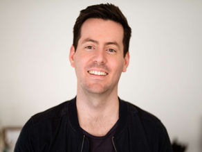 Smiling person in a black jacket against a neutral indoor background, conveying a friendly mood.