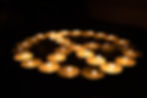 Small tea light candles are all lit and arranged to make a peace symbol against a dark background 