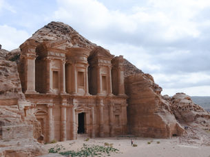 A wide shot of the Monastery (Ad-Deir) in Petra, Jordan. The massive, ancient structure with a classical facade of columns and pediments is carved directly into the face of a reddish-brown mountain. The sky above is filled with soft, grey clouds, and the surrounding terrain is rocky and arid.