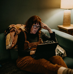 A woman on a couch, looking tired and frustrated while intensely focused on her laptop, hand to her head in exasperation.