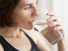 Woman drinking water from a clear glass, wearing a black tank top. Neutral background with soft lighting. Focus on hydration and tranquility.
