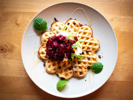 Goldbraune herzförmige Waffel auf weißem Teller mit Beerenkompott, Joghurt und Minzblättern – frisch serviert auf Holztisch.