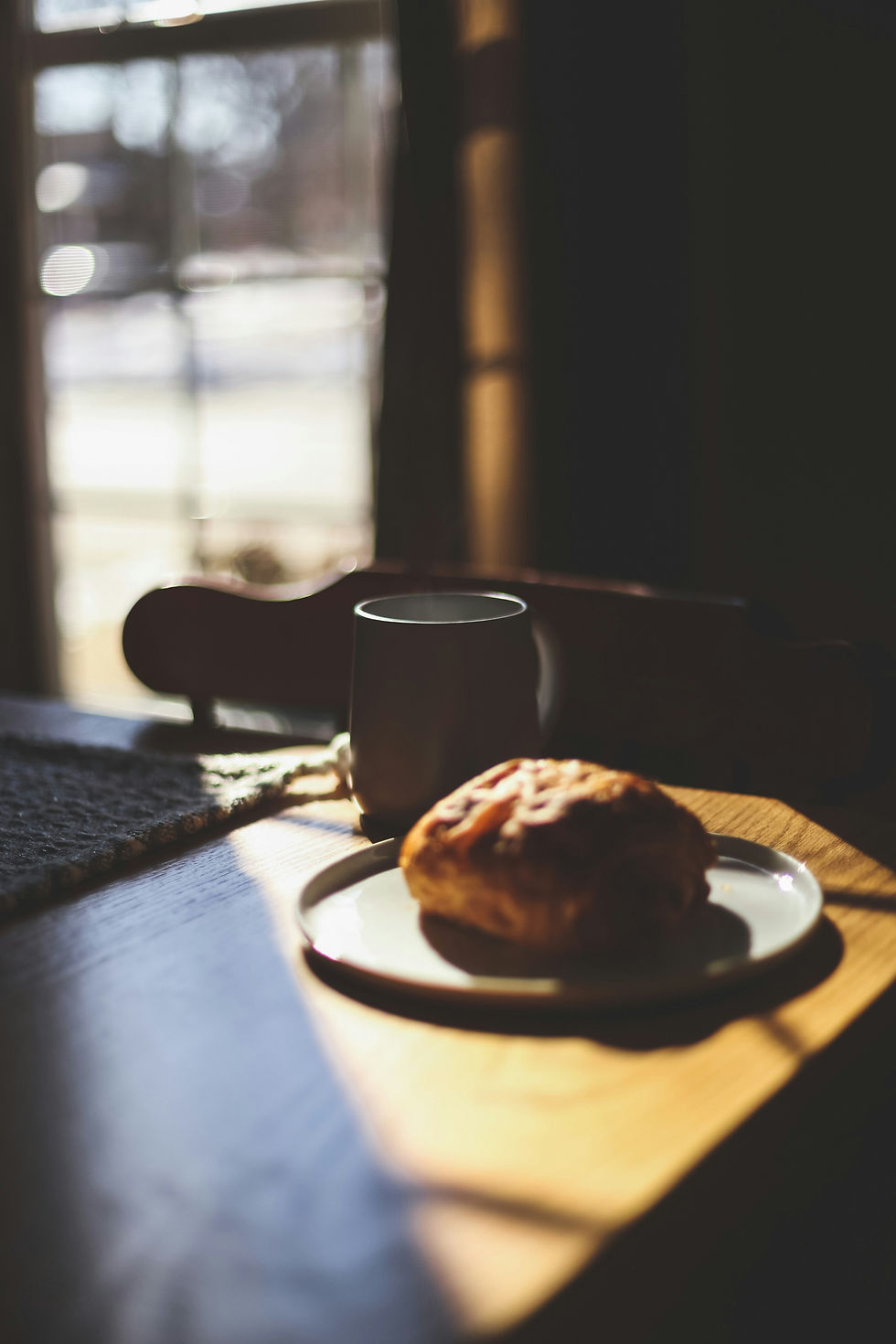 Pastry on a plate and mug on a sunlit wooden table. Soft, warm morning light through a window creates a cozy, calm atmosphere.