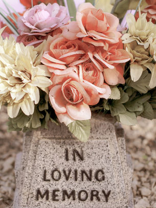 Bouquet of pink and cream flowers on a stone marker with "IN LOVING MEMORY" text, set on a gravel background. Gentle, somber mood.