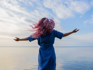 Person with pink hair and blue dress stands with arms outstretched facing a calm sea. Sky is partly cloudy, evoking freedom and serenity.