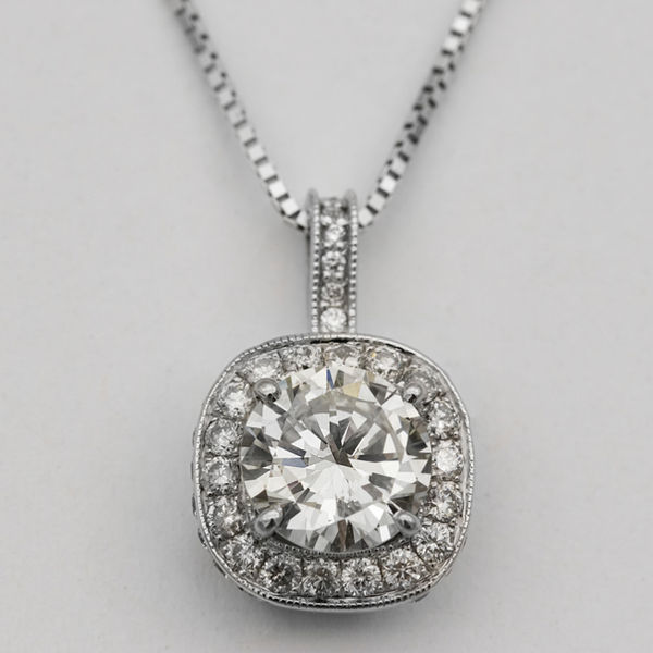 A single hook diamond pendant featuring a large solitaire diamond with a halo of small round brilliant cut diamonds, in white gold, and attached to a white gold chain.