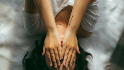 Person in white lying on a fluffy rug, covering face with hands, wearing rings. The mood is calm and introspective, with soft lighting.