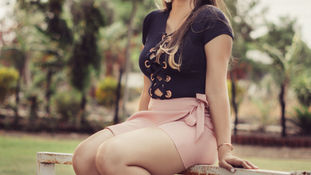 Woman in black top and pink shorts sits joyfully on a white fence in a grassy area with trees. She wears white sneakers and earrings.