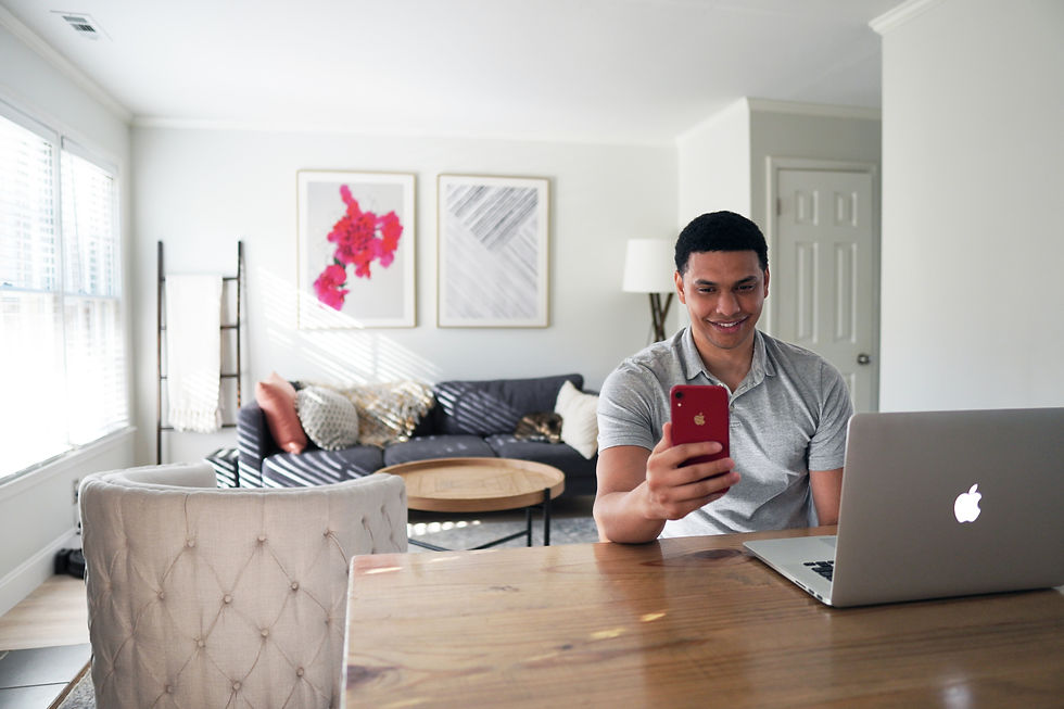 Man smiling at phone, sitting at wooden table with laptop. Bright living room, colorful art on wall, sofa, and natural light streaming in.