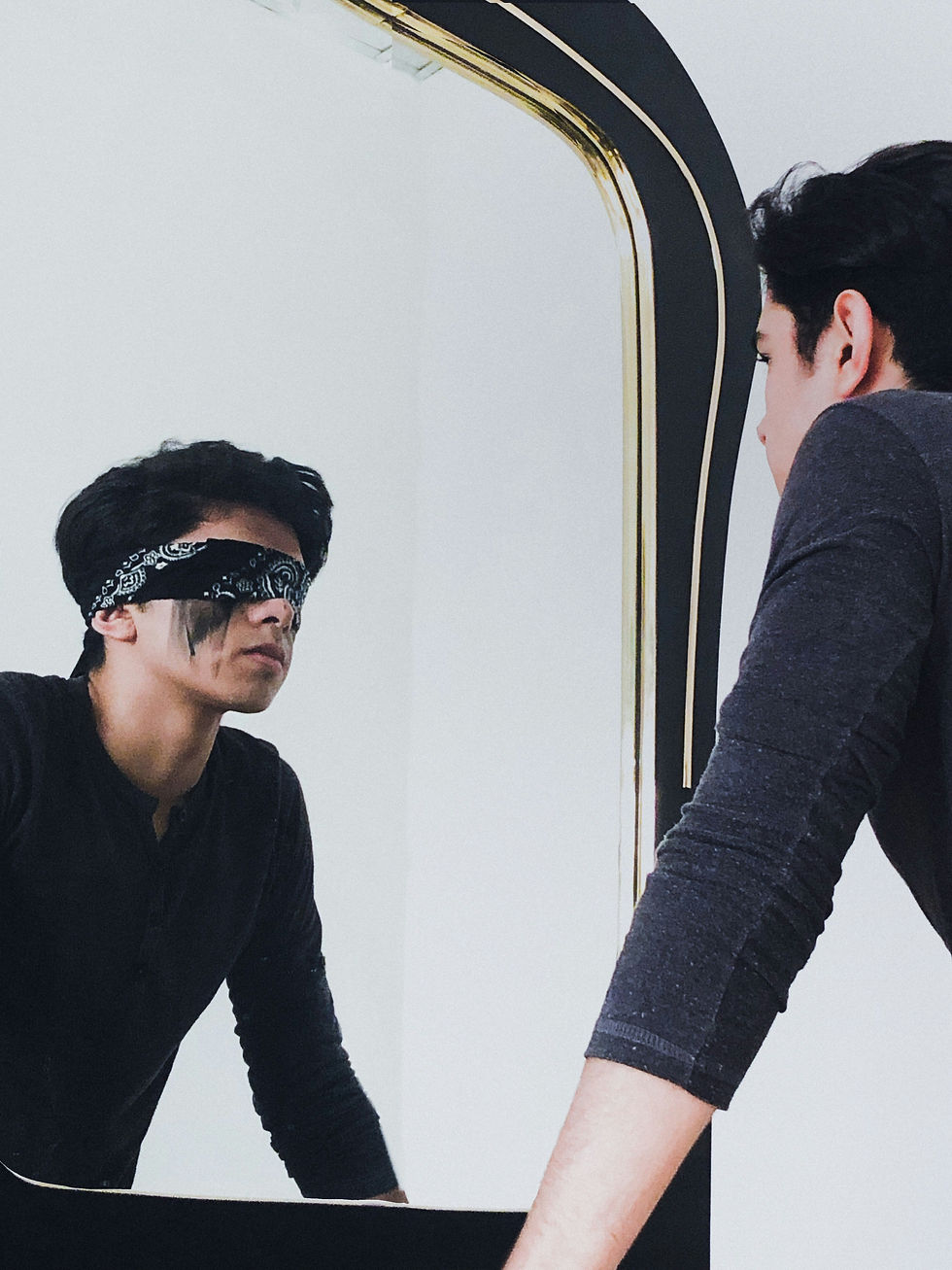 A man stands in front of a mirror, wearing a black bandana over his eyes with black paint on his face, reflecting a contemplative mood.