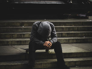 Dreary background with a person sitting on concrete outdoor steps, on a skateboard, wearing a gray hoodie with hood on, head looking down into lap with hands together