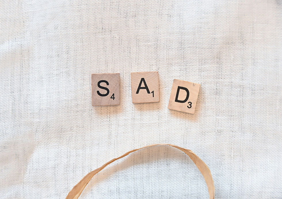 Scrabble tiles spell "SAD" on textured white fabric, above a curved brown ribbon, creating a sad face impression.