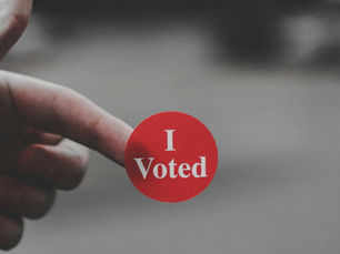 A finger points at a red I Voted sticker against a blurred gray background.