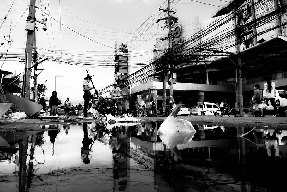 街中の白黒写真。人々が水たまりの周りを歩き、電線が複雑に絡まる背景。遠くにビルや車が見える。沈んだ雰囲気。