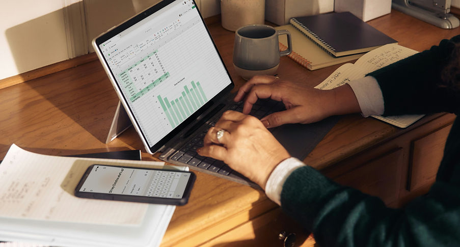 Person arbejder ved skrivebord med laptop, der viser dataanalyse, og mobiltelefon liggende ved siden af