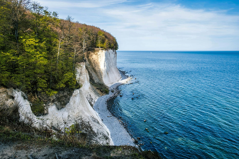 White chalk cliffs and green trees above a calm blue sea, with a pebble shore and distant sailboat.