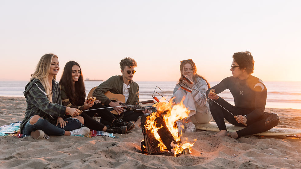 Group of young adults sitting around a beach bonfire at sunset, smiling and socializing.
