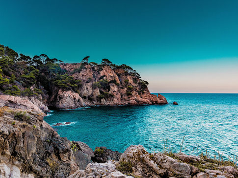 Rocky, tree covered cliffs jut out into the calm blue sea on the coast of Spain.