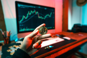 Hand holds bitcoin coin in front of a computer screen displaying a rising stock chart. Red and teal lighting creates a dynamic mood.