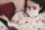 Woman wearing a mask reads a book while lying on a floral quilt. Soft lighting and a calm, focused mood.