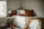 Cozy bedroom with a white bed, dinosaur pillow, and brown throw. A side table holds books and a lamp. Window light adds warmth.