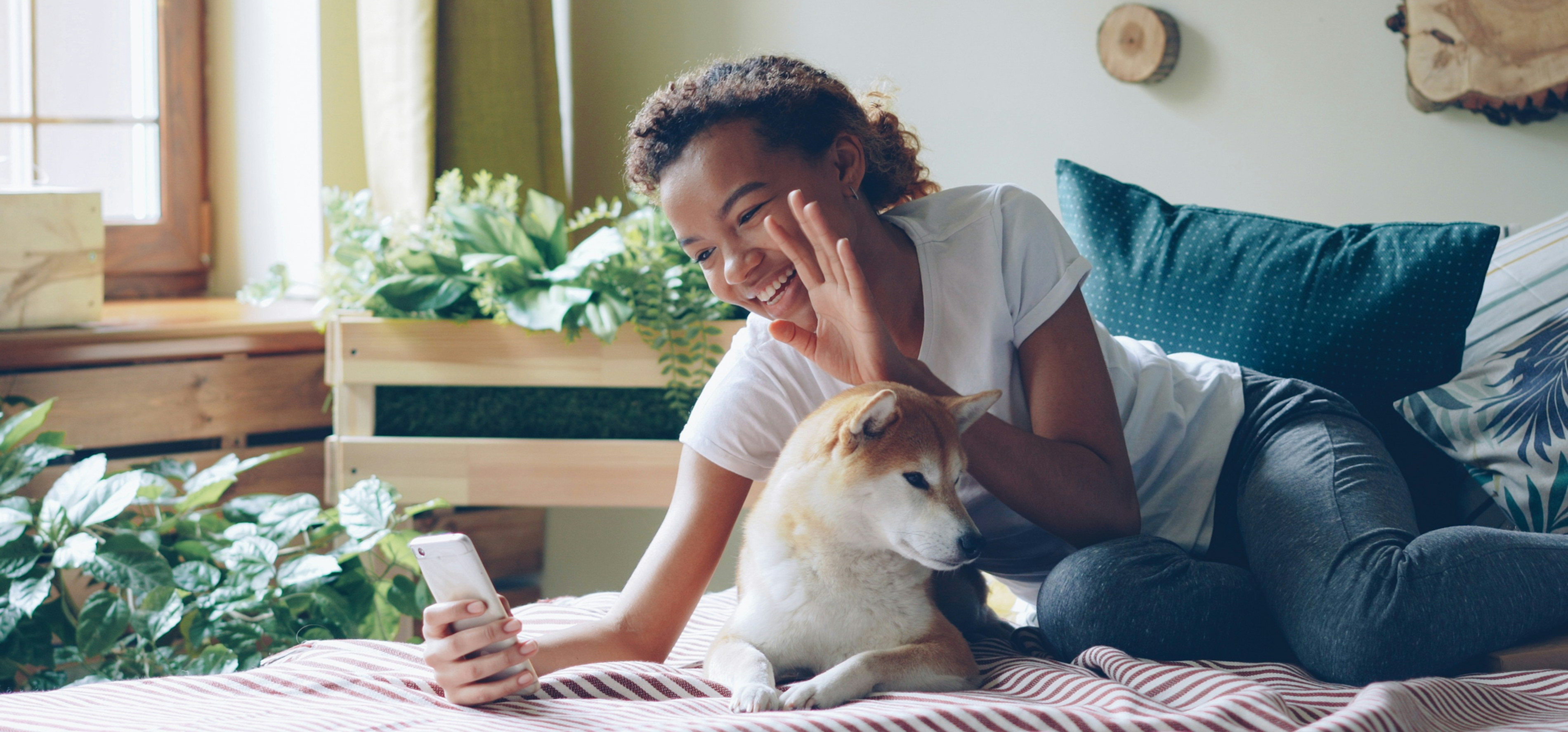 Smiling woman on a bed takes a selfie while cuddling a Shiba Inu in a cozy sunlit room with plants and wood decor